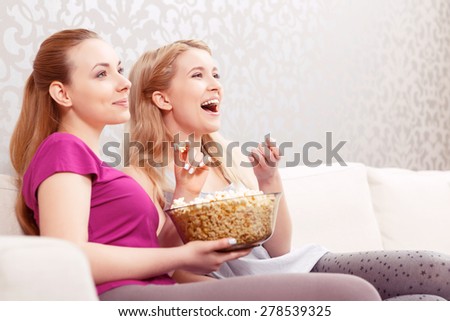 Comedy movie. Two young beautiful girls sitting on a white couch watching a movie and laughing while eating popcorn at pajama party side view