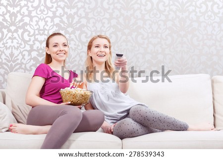 Movie night. Two young beautiful girls sitting on a white couch watching a movie while holding remote control and eating popcorn at pajama party full length