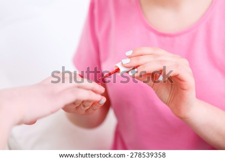 Close up hands of a young girl wearing pink t-short and painting nails of her friend in red color at pajama party in the light room