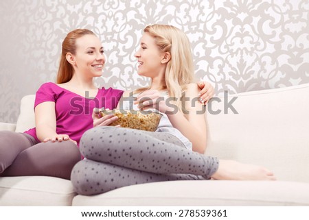 Movie night. Two young beautiful girls sitting on a white couch discussing a movie while eating popcorn at pajama party full length
