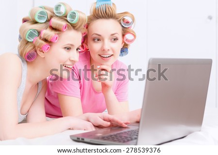 Young beautiful girls wearing pajamas and colorful hair rollers lying on the bed and looking interestedly at laptop at home party in the light room