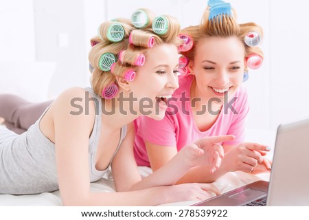 Young beautiful girls wearing pajamas and colorful hair rollers lying on the bed laughing and looking at laptop at home party in the light room
