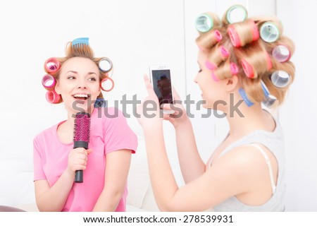 Sing a song. Two happy young friends having fun making mobile photos and pretending singing into the hairbrush wearing pajamas and colorful hair curlers at home