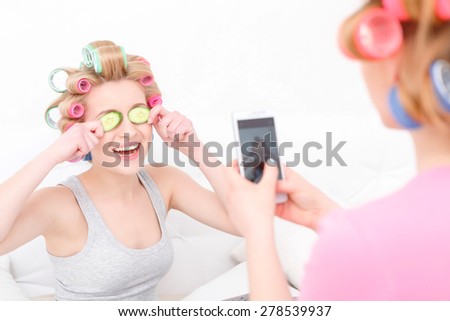 Pajama party. Young blond girl making mobile photo of her beautiful friend laughing and holding cucumbers on her eyes wearing colorful hair curlers and grey shirt