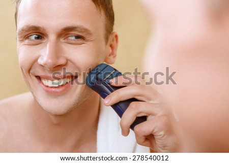 Using technologies. Young handsome man shaving with help of electric razor in front of mirror n bathroom.