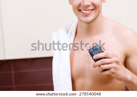 Going to start. Close up of smiling topless man with towel on his shoulder holding electric razor