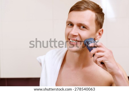 Clean shave. Smiling young handsome man shaving with help of electric razor in front of mirror n bathroom.