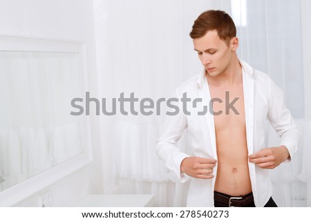 Sensory perception. Handsome muscular man holding shirt that he is wearing against background of white decorated room