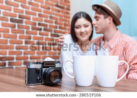 Table with two cups and a vintage camera standing on it, sweet couple of asian girl and her boyfriend wearing a hat on a background
