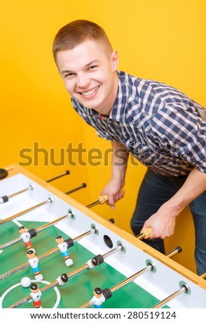 Portrait of a young smiling boy standing near table hockey and holding the handles wearing jeans and checkered shirt yellow wall on a background