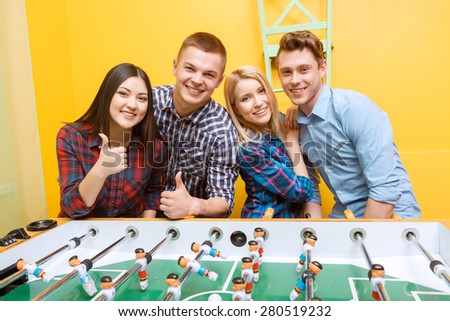 Double date. Two happy young pairs standing smiling near table hockey wearing casual holding thumbs up clothes in a yellow room