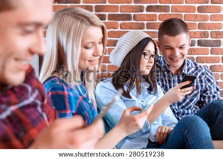 Young happy students sitting in a row using their smartphones while meeting in a cafe with their friends wearing casual clothes, brick wall on the background