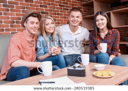 Friendship. The company of young happy friends sitting together on a couch in a cafe, smiling and holding cups of tea and coffee , brick wall on the background