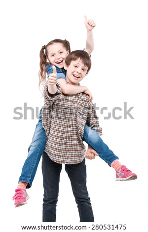 Be my horse. Small handsome boy holding his small pretty sister on his back while both are smiling and showing thumbs up isolated on white background