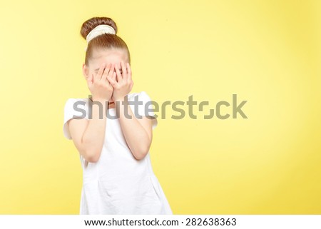 Portrait of a small pretty girl standing and holding her hands on her face hiding it completely wearing a white t-shirt with big bun hairstyle, isolated on a yellow background