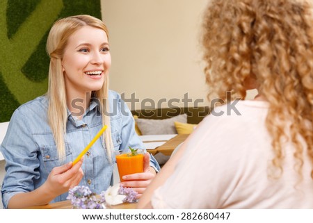 Interesting news. Beautiful smiling blond girl sitting in front of another woman with curly hair in cafe, holding straw and glass of cocktail