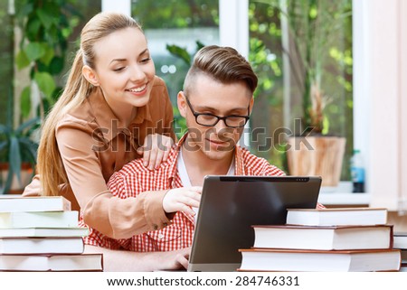 Portrait of a handsome smart student wearing red checkered shirt and glasses working with a laptop and a beautiful girl standing behind him helping and smiling, a stack of books on the table
