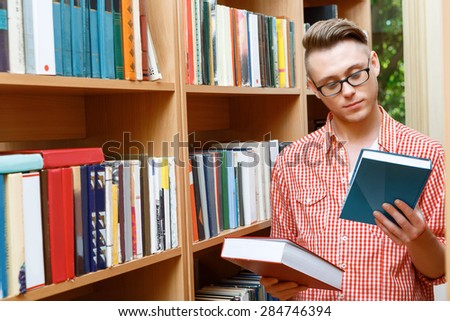 Portrait of a handsome smart student wearing red checkered shirt and glasses standing near a bookshelf in a library, holding two books in his hands and choosing, selective focus
