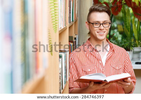 Portrait of a handsome smart student wearing red checkered shirt and glasses standing near a bookshelf in a library, holding a book in his hands and smiling, selective focus