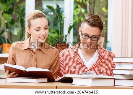 Portrait of a handsome smart student wearing red checkered shirt and glasses reading a book and his beautiful friend looking in his book smiling, a stack of books on the table