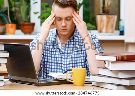 Portrait of a handsome smart student wearing glasses and blue checkered shirt sitting at the table in library thinking and holding hands on temples closing his eyes, a stack of books on the table