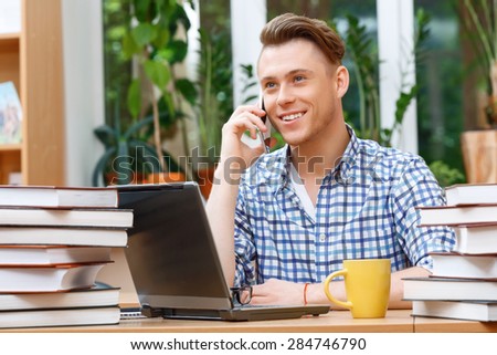 Portrait of a handsome smart student wearing glasses and blue checkered shirt sitting at the table in library talking on a phone smiling, a stack of books laptop and yellow cup on the table