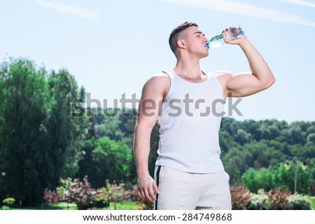 Portrait of a young handsome muscular sportsman posing, drinking water from the bottle tired of exercises, wearing white sportswear full length in the park