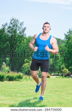 Portrait of a young handsome muscular sportsman running on a green grass smiling and listening to music, wearing black blue sportswear during training full length in the park