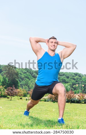 Portrait of a young handsome muscular sportsman leaning on left leg and holding his hands crossed behind his head and smiling, wearing black blue sportswear during training in the park full length
