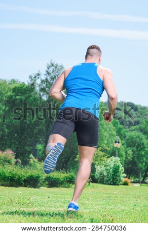 Portrait of a young handsome muscular sportsman running on a green grass back view, wearing black blue sportswear during training full length in the park