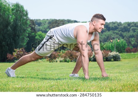 Portrait of a young handsome sportsman standing in a pose ready to run and smiling, wearing white sportswear making body exercises full length in the park