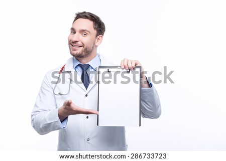Look here. Smiling doctor holding the sheet of paper and pointing it with his hand while standing isolated on white background.