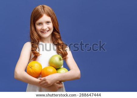 Real emotions. Vivacious young red-haired girl keeping apples and oranges in her hands while expressing positivity on blue background.