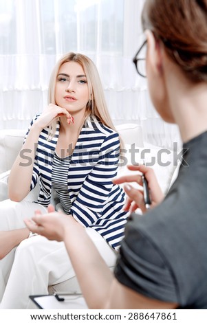 Young blond woman sitting on a couch propping her head telling about her problems, doctor with clipboard listening to them and making notes during therapy session, selective focus
