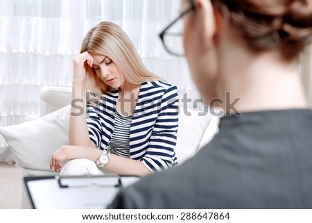 Young blond woman sitting on a couch holding her head sadly crossed telling about her problems, doctor with clipboard listening to them and making notes during therapy session, selective focus