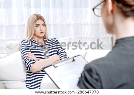 Young blond woman sitting on a couch with her arms crossed telling about her problems, doctor with clipboard listening to them and making notes during therapy session, selective focus