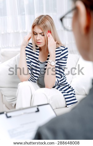 Young blond woman sitting on a couch holding her head sadly crossed telling about her problems, doctor with clipboard listening to them and making notes during therapy session, selective focus