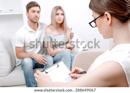 Young couple sitting on a couch at psychologist hugging telling about their relationships, female doctor with clipboard in hands listening to them and making notes, selective focus