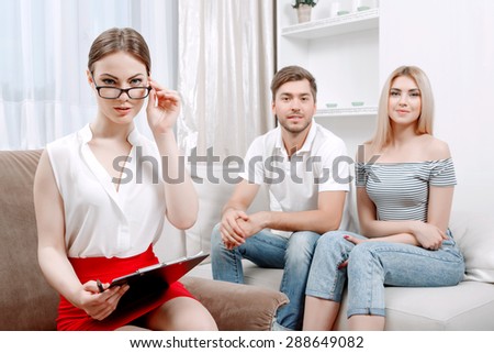 Portrait of a beautiful psychologist wearing white blouse and black skirt touching her glasses sitting at her office during therapy session, patients young couple on a couch smiling, selective focus