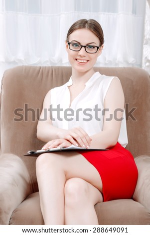 Portrait of a professional female psychologist wearing white blouse red skirt and glasses, sitting smiling in the armchair holding a clipboard in her office during therapy session