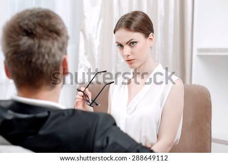 Portrait of a professional female psychologist wearing white blouse holding glasses in her hand and looking seriously at her client sitting in her office during therapy session, selective focus