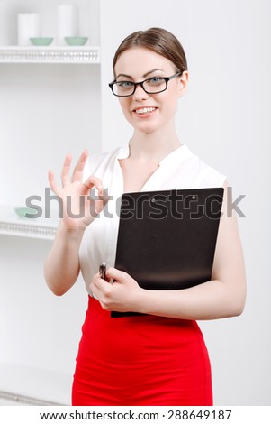 Portrait of a professional female psychologist wearing white blouse and red skirt and glasses, standing smiling holding a clipboard showing everything is good, in her office during therapy session
