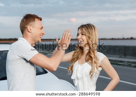 Waist up portrait of a handsome young man and beautiful blond girlfriend with curvy hair standing near their white car giving high five and smiling, stunning landscape on the background