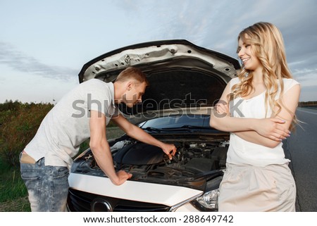 Car trip. Portrait of a beautiful blond girl with curvy hair standing watching her handsome boyfriend fixing something broken in their car, stunning landscape on the background