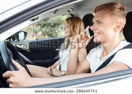 Sweet drive. Portrait of a handsome young man driving a white car and talking on his mobile phone while his beautiful blond girlfriend with curvy hair sitting near smiling happily