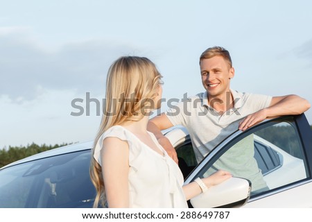 Sweet couple. Portrait of a beautiful blond girl with curvy hair looking back at her handsome boyfriend standing near his white car, stunning landscape on the background