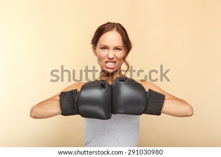 Portrait of a beautiful young woman with brown hair wearing grey top and black boxing gloves looking aggressive holding her hands together in front of her body, isolated on beige background