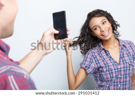 Back view of a young man making a photo of his charming mulatto girlfriend posing for him and smiling, isolated on grey background, selective focus