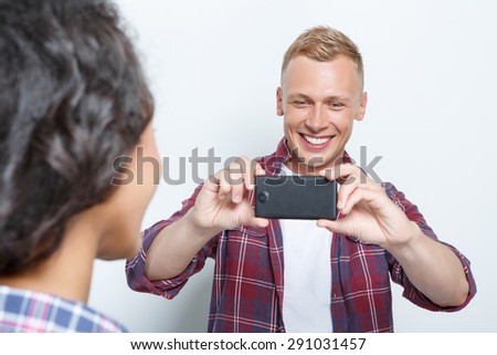 Back view of a brunette girl posing and a blond happy smiling young man making a photo on his smartphone, isolated on grey background, selective focus