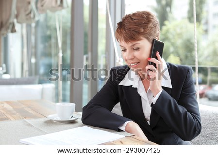 Portrait of a beautiful businesswoman wearing formal suit sitting at the table looking at some documents in front of her and talking on her mobile phone, in a restaurant during business lunch